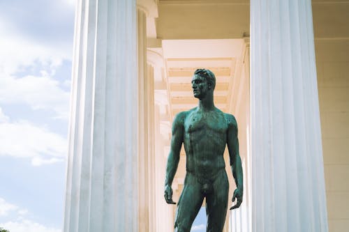 Free From below of statue of nude man standing between white marble columns of historic building in sunny day Stock Photo