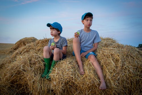 Two Boys Sitting on a Haystack while Wearing a Cap