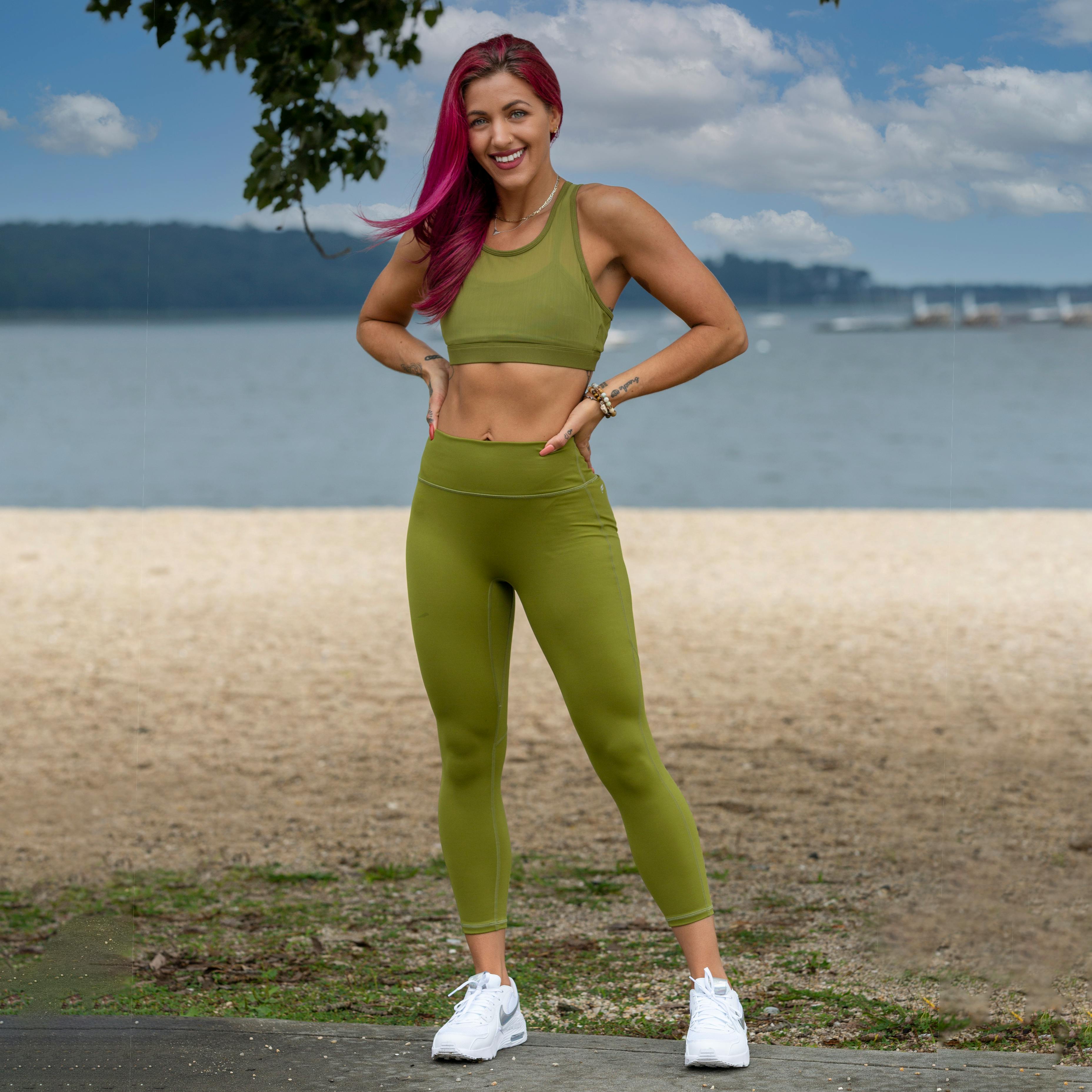 Attractive sporty woman wearing green top and gray leggings standing near  green background holding pink dumbbells and smiling. Stock Photo