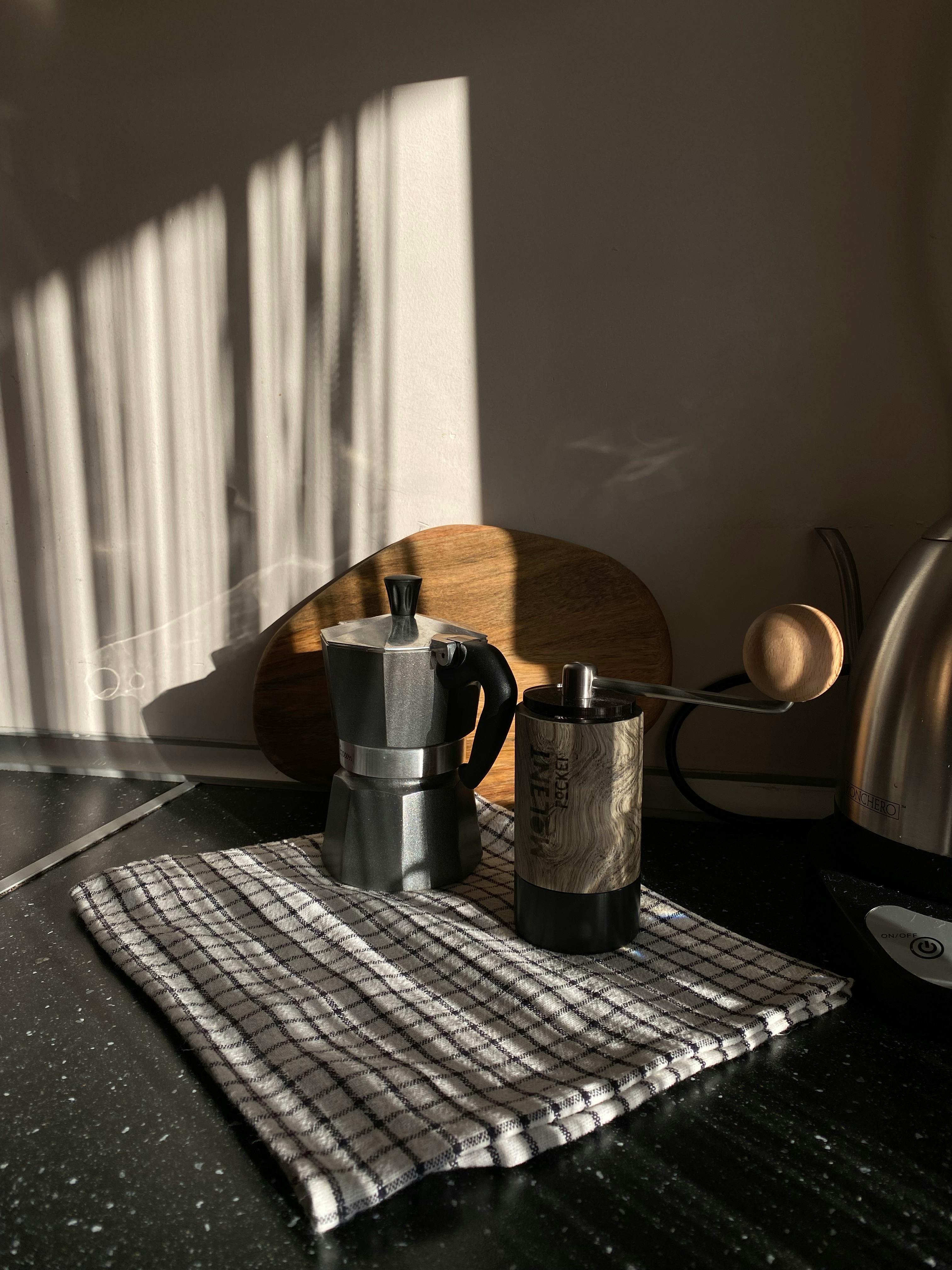 A warm kitchen setting with a moka pot and coffee grinder on a towel, bathed in sunlight.