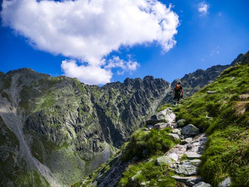 Free A hiker climbs a rocky trail in a scenic mountainous landscape, under a bright blue sky with scattered clouds. Stock Photo
