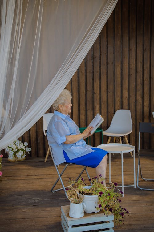 An Elderly Woman Sitting on a Folding Chair while Reading a Book