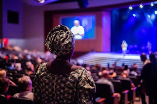 A woman in traditional African attire attending a public speaking event in a large auditorium.