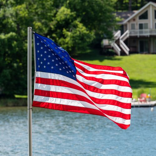 Free Vibrant American flag waving by a picturesque lake with a house backdrop, capturing summer spirit. Stock Photo