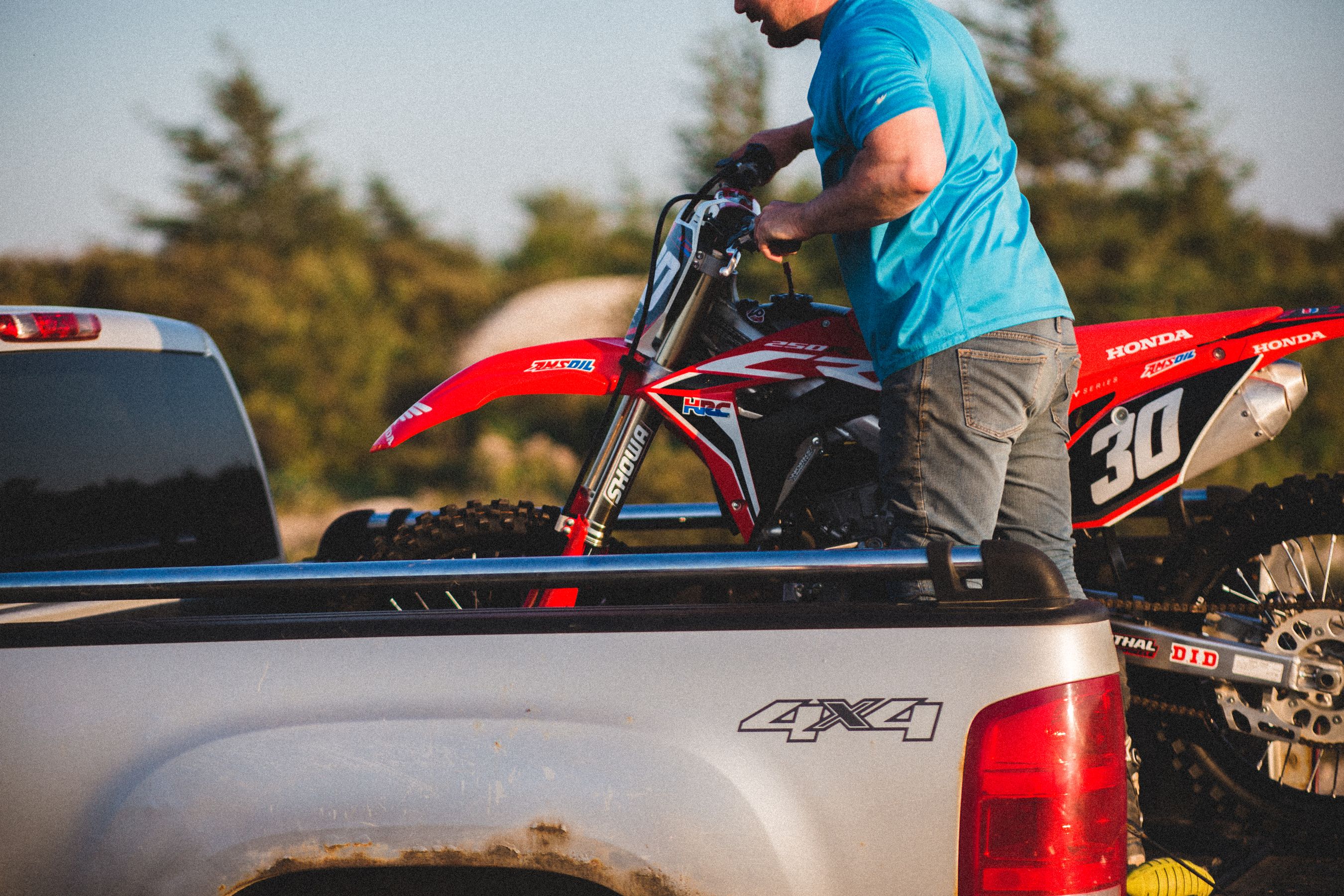 Man loading a red dirt bike into a silver pickup truck bed at golden hour before tying it down for transport