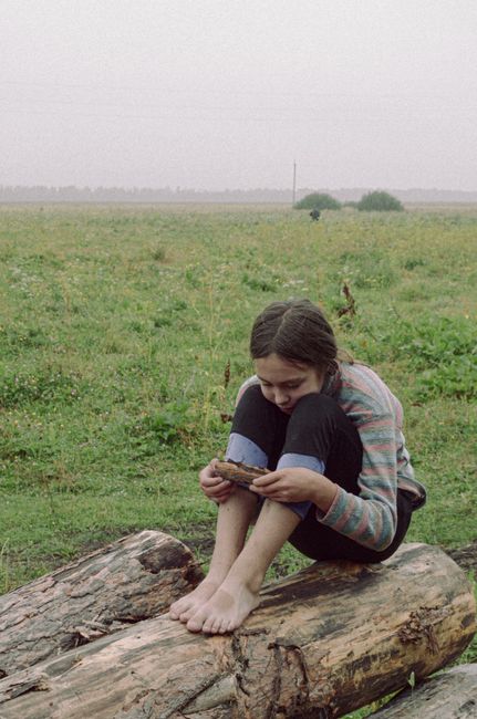 A teenage girl sits barefoot on a log in a foggy field, holding bread.