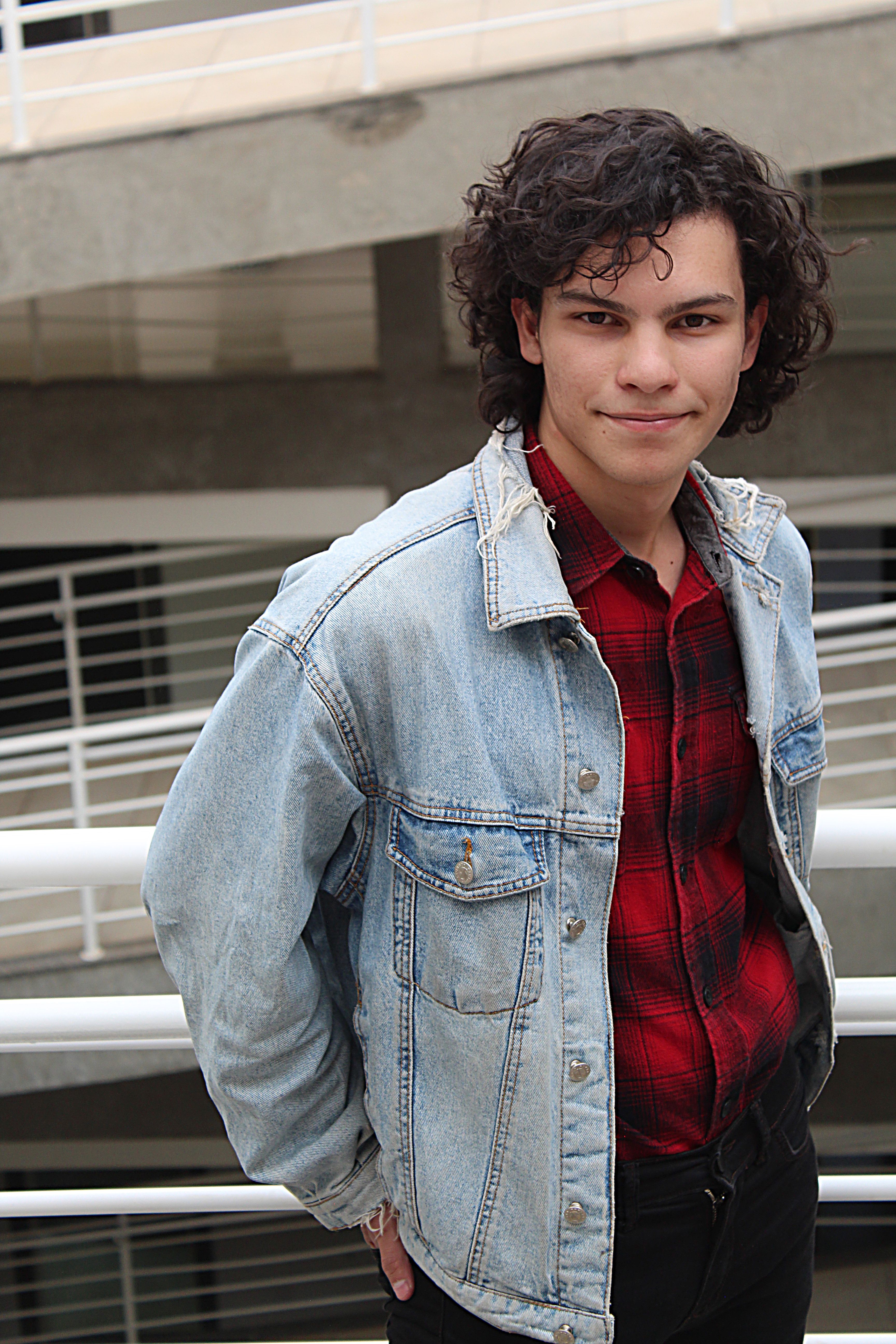 A young man in a denim jacket poses confidently outdoors, showcasing his casual style.