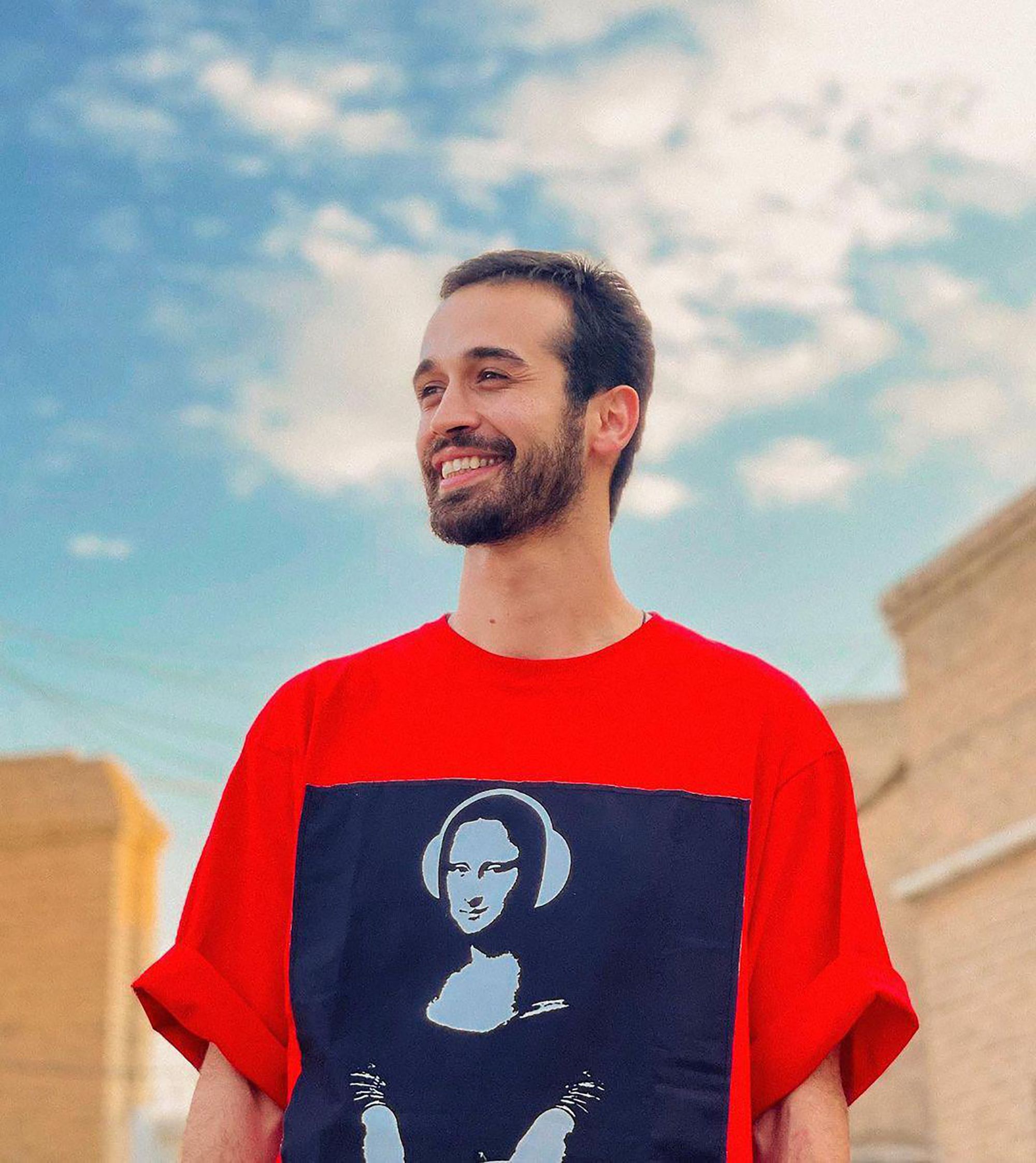 A bearded man smiling outside wearing a modern red shirt with a printed design.