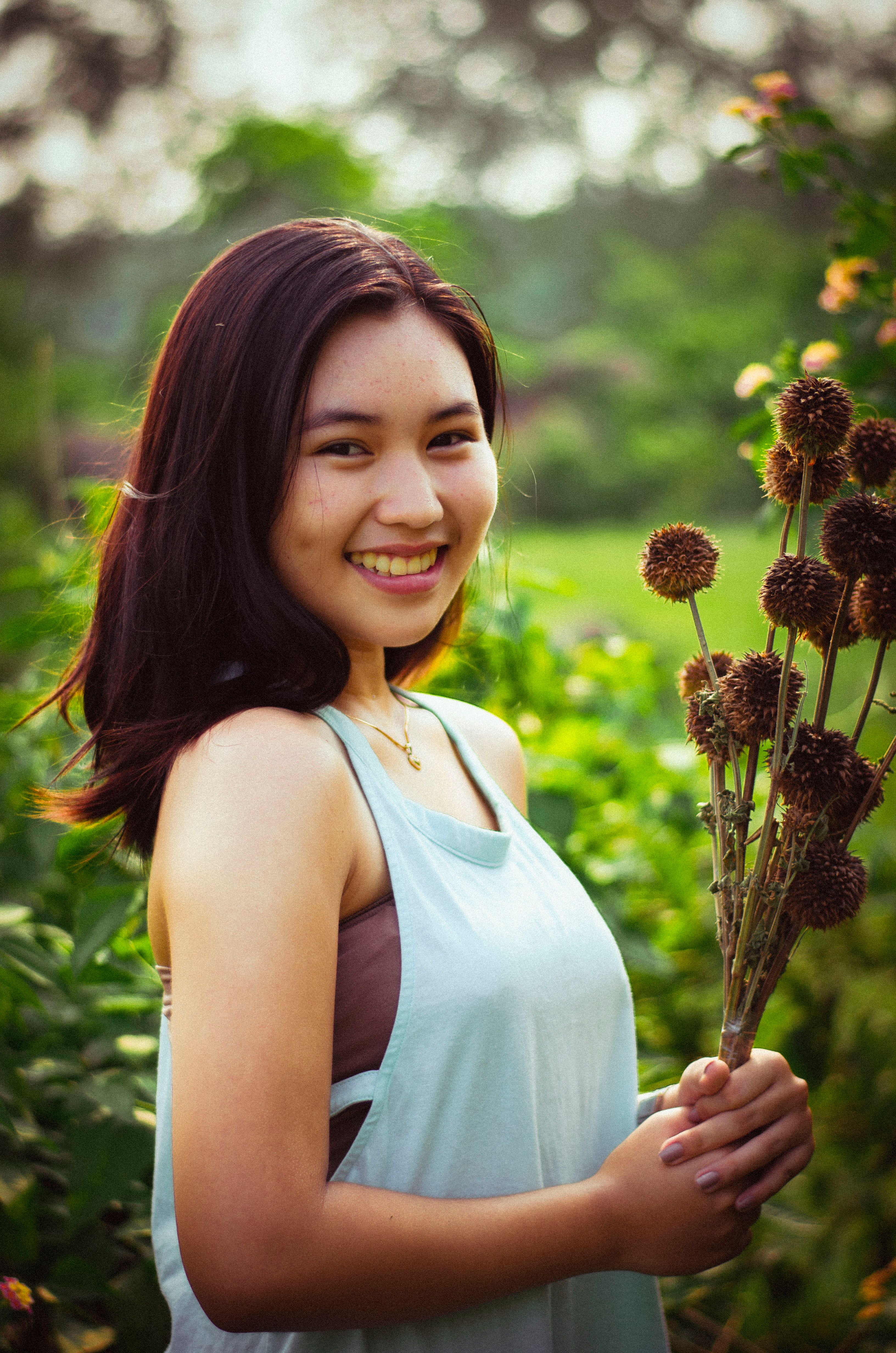 Smiling woman holding dried flowers in a garden setting on a sunny day.
