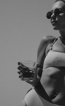 Black and white photo of a woman in swimwear holding a drink, wearing sunglasses, exuding a fashionable vibe.