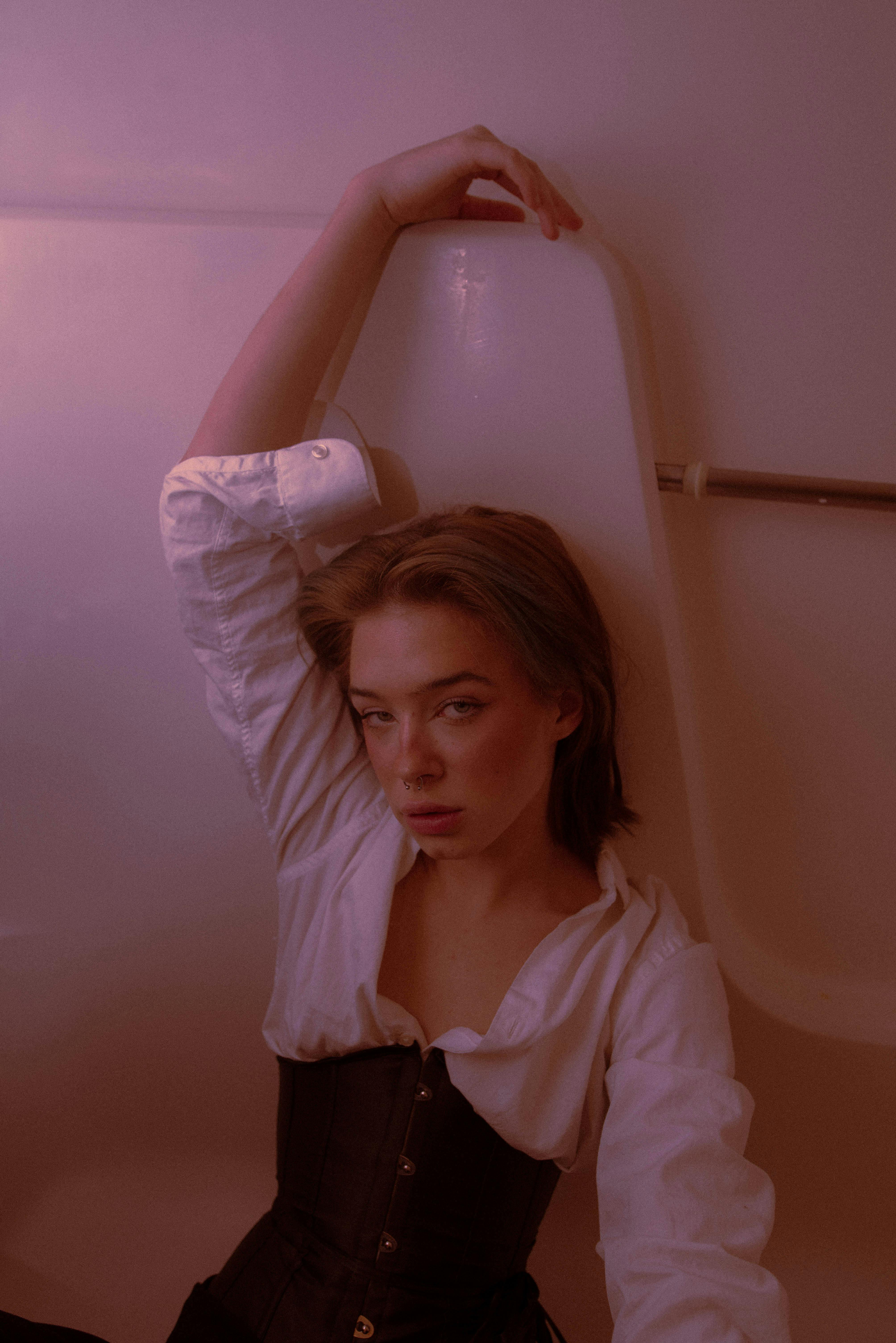 Elegant woman in white shirt and corset posing indoors with arm raised, in soft lighting.