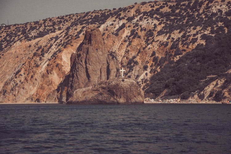 White Cross Symbol On Top Of The Rock Formation Near Body Of Water