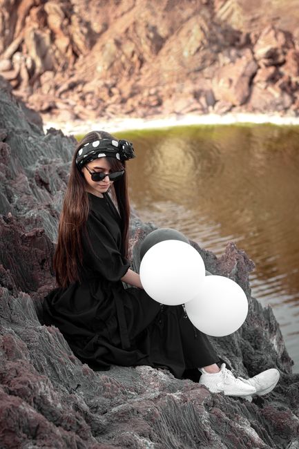 A stylish woman in sunglasses sits on rocks holding balloons by a lake in Tehran.