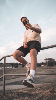 Young man in casual attire sitting on railing outdoors using smartphone on a sunny day.