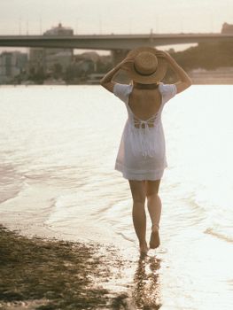 A woman in a white dress and sun hat walks barefoot on a sandy beach at sunset.