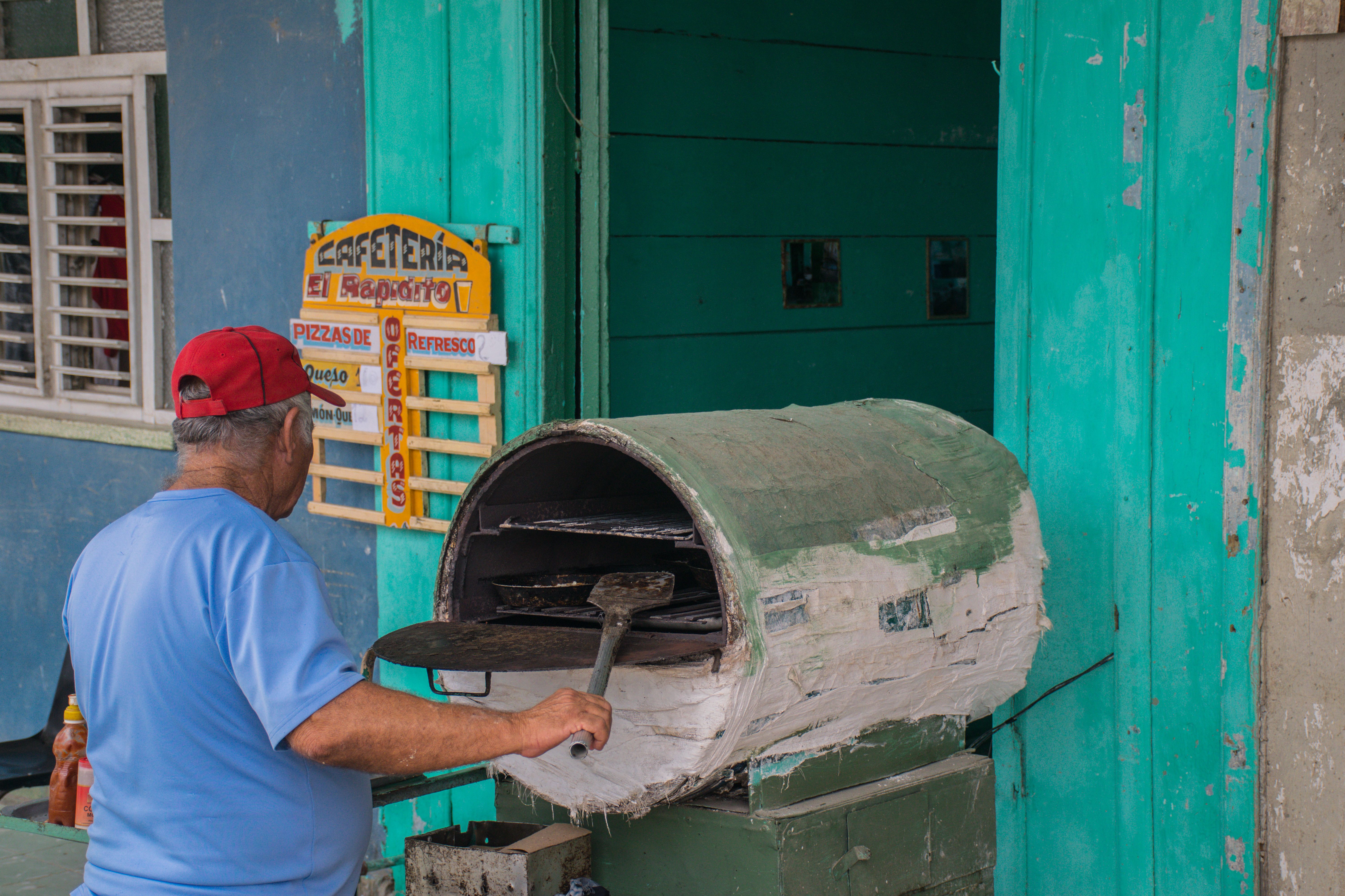 An elderly man in a blue shirt cooks with a makeshift oven outside a colorful building in Cuba.