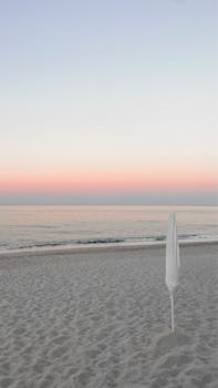 Peaceful beach scene at sunset with pastel sky and closed umbrella.