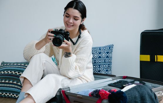 Woman sitting by an open suitcase indoors, holding a camera and smiling.