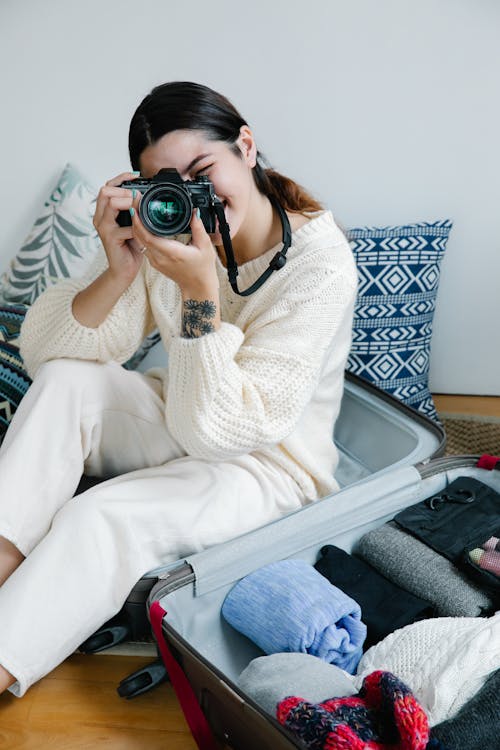 Free A Woman in White Sweater Sitting on Her Luggage while Holding a Camera Stock Photo