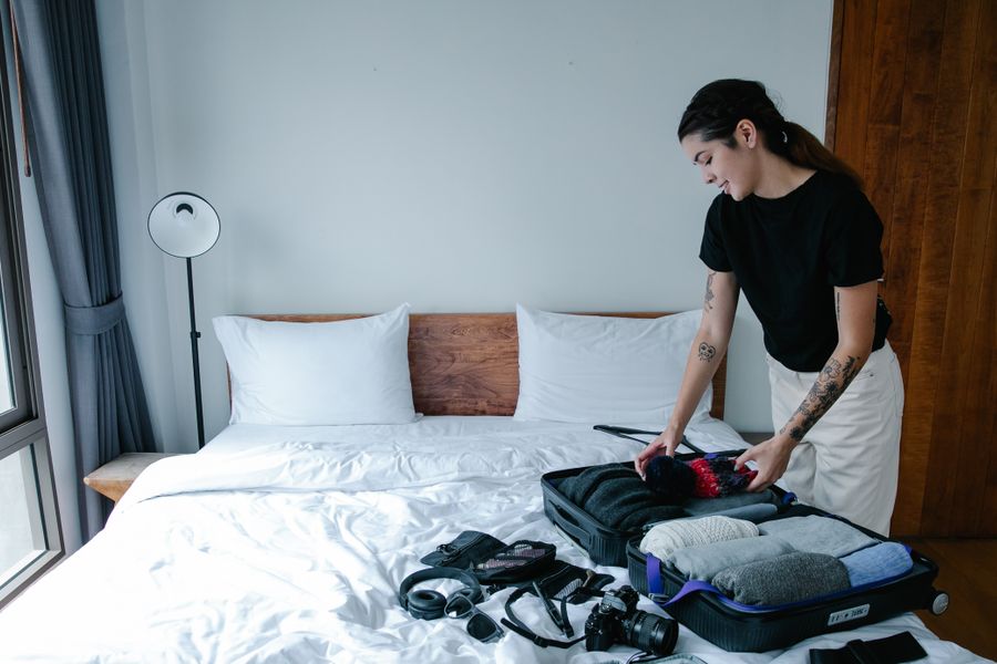 Woman packing suitcase with clothes and camera in a bright bedroom.
