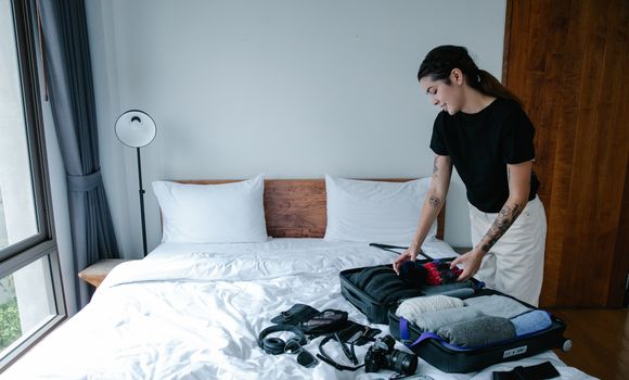Woman packing suitcase with clothes and camera in a bright bedroom.