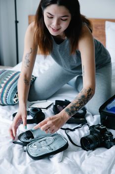 Smiling woman with tattoos organizing camera equipment on bed indoors.