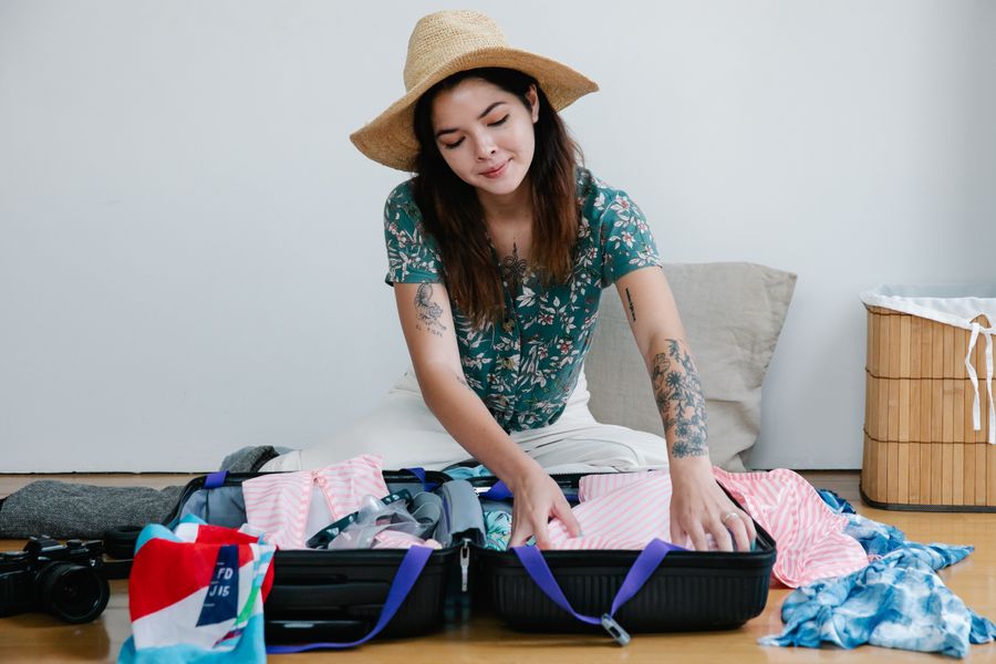 Woman packing suitcase with clothes for a vacation, wearing straw hat and smiling.