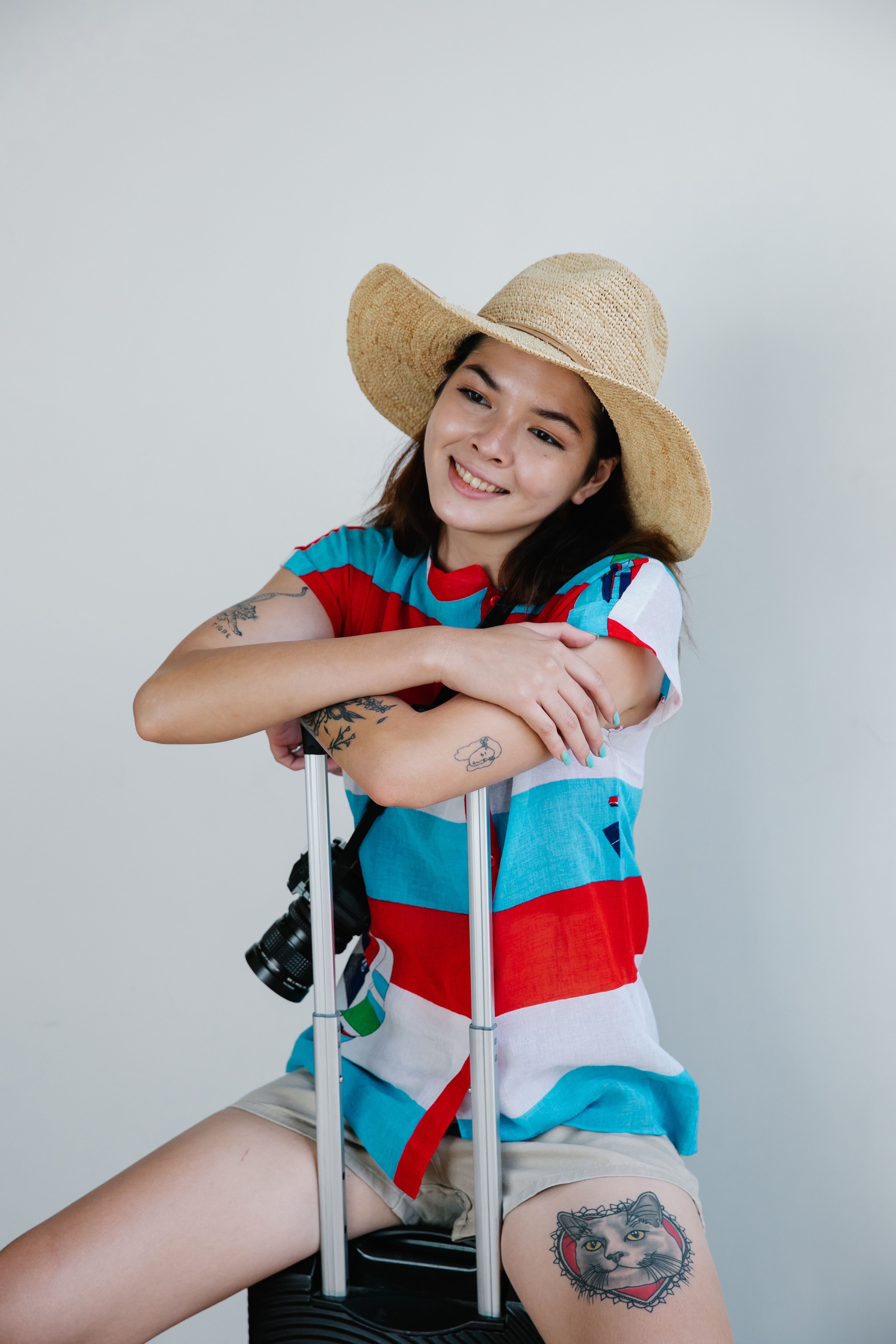 Smiling Asian woman with tattoos and straw hat, sitting with luggage in studio.