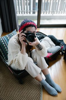 Young woman in a cozy sweater and beanie sitting on luggage, holding a camera indoors.