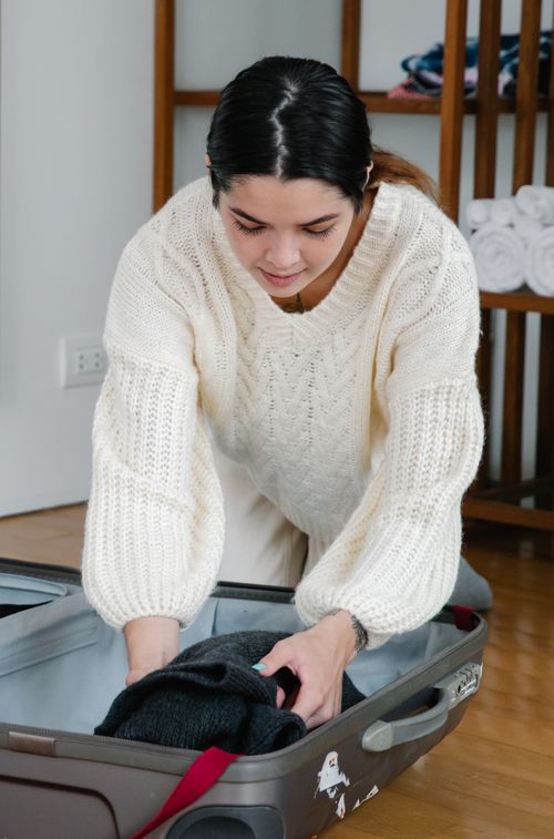 Free Woman in cozy sweater packing clothes into a suitcase, preparing for travel. Stock Photo