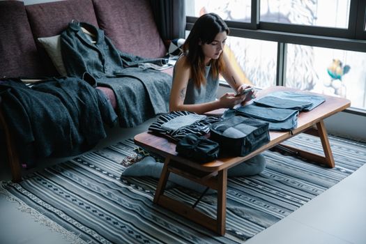 Young woman sitting on floor packing clothes, using smartphone in bright indoor setting.