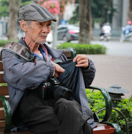 Free Senior man with a camera sitting on a park bench, reaching into his pocket. Stock Photo