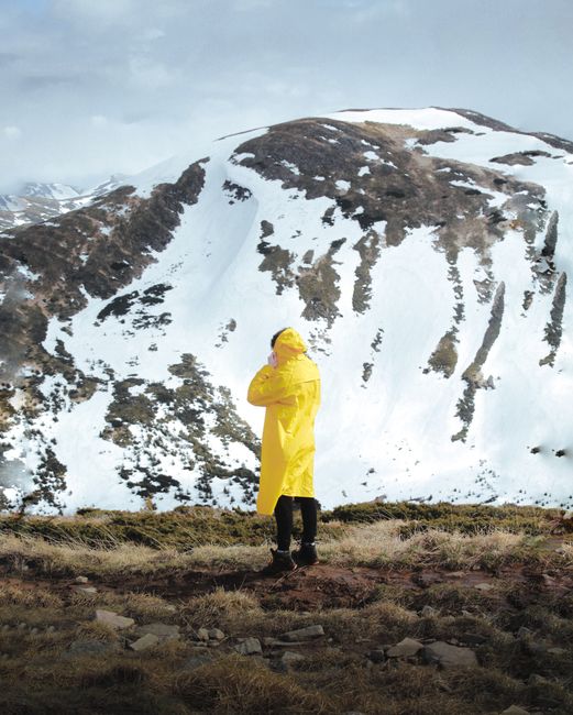 A person in a yellow raincoat stands on a mountain summit, facing snow-capped peaks.