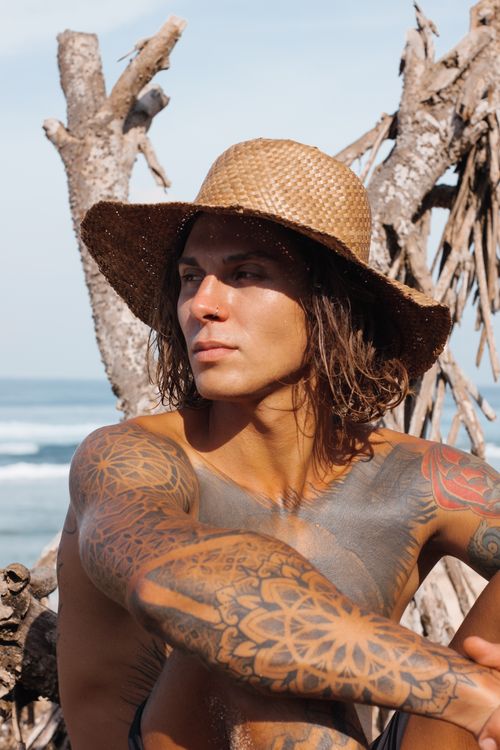 Free Shirtless tattooed man in a straw hat outdoors by the beach, looking thoughtful. Stock Photo