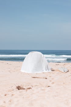 A peaceful beach scene with a tent and surfboard against the ocean waves under a clear sky.