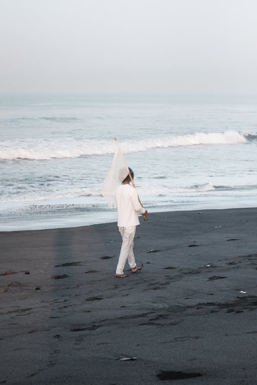 Woman in White Pullover and Pants Walking in Beach with Flag
