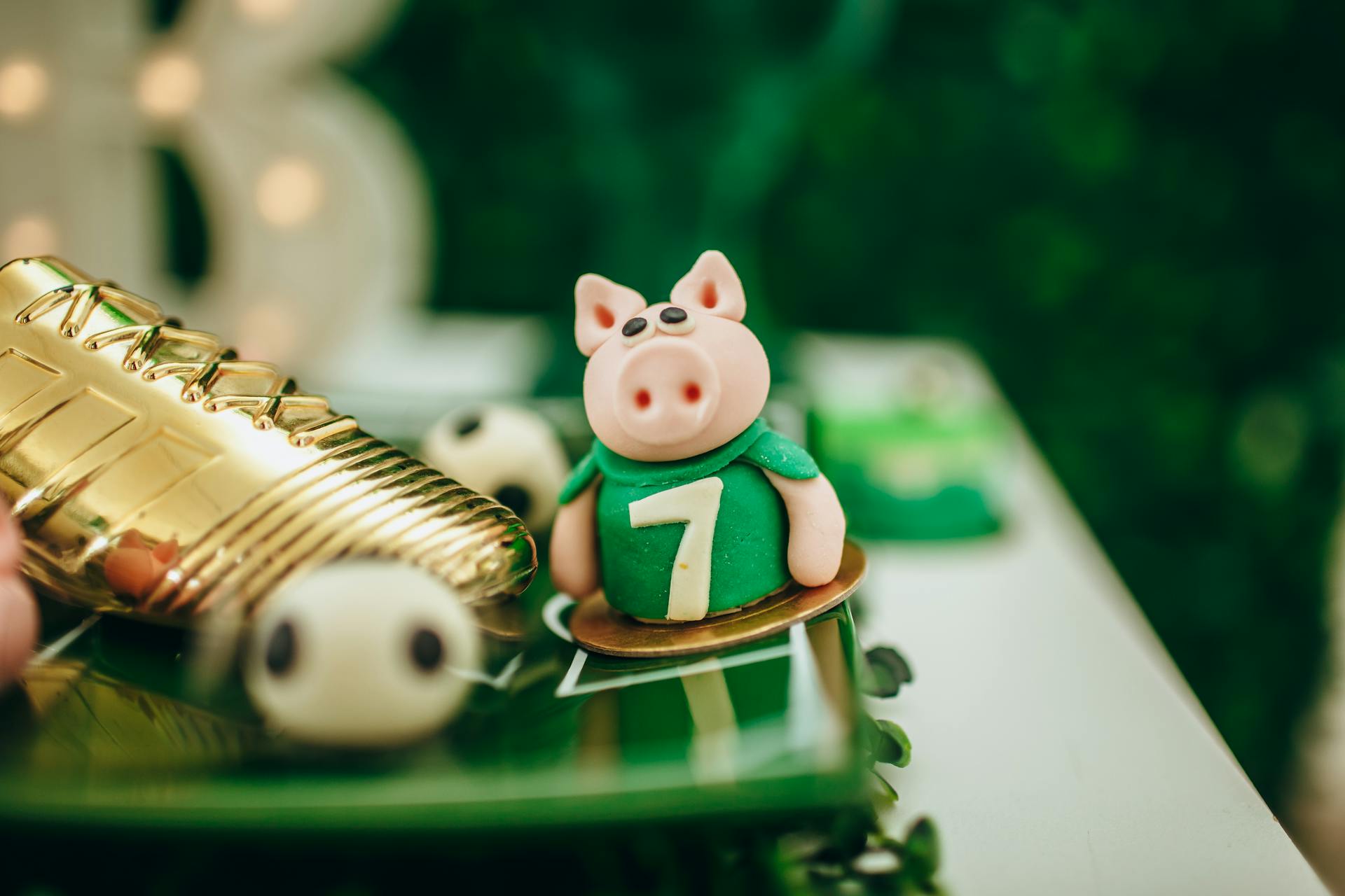 A close-up of a toy pig cake topper wearing a football jersey number 7, beside a golden football boot.