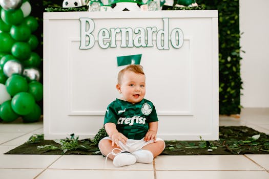 Cute baby boy in a sports jersey sitting indoors, surrounded by colorful decorations. Ideal for birthday or celebration themes.