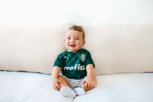 Cute baby smiling on a sofa, wearing a green sports jersey indoors.