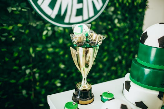 A golden trophy on a decorated table with soccer-themed cake and greenery backdrop.