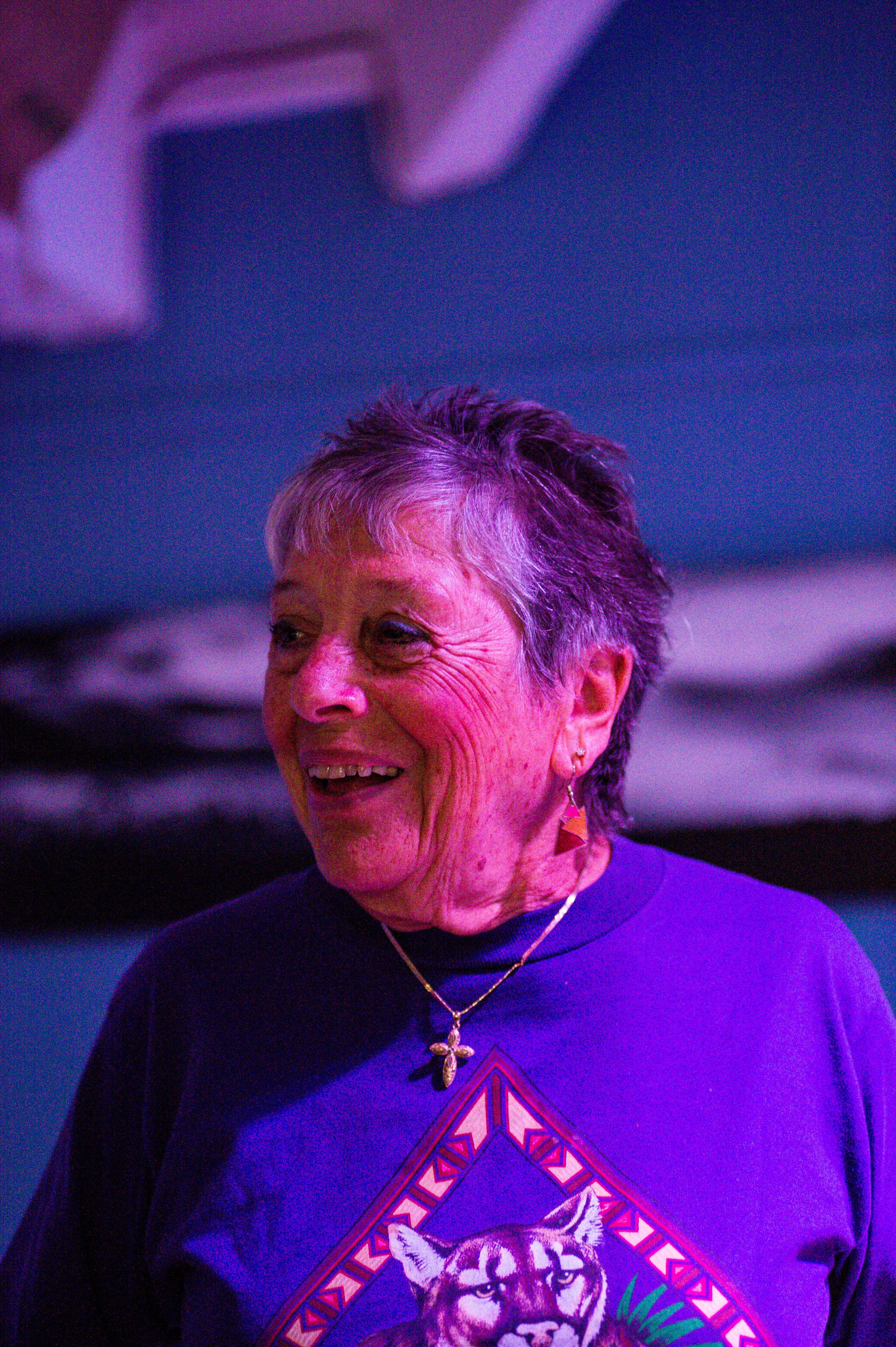 Elderly woman in a purple shirt smiling warmly, captured indoors with a colorful background.
