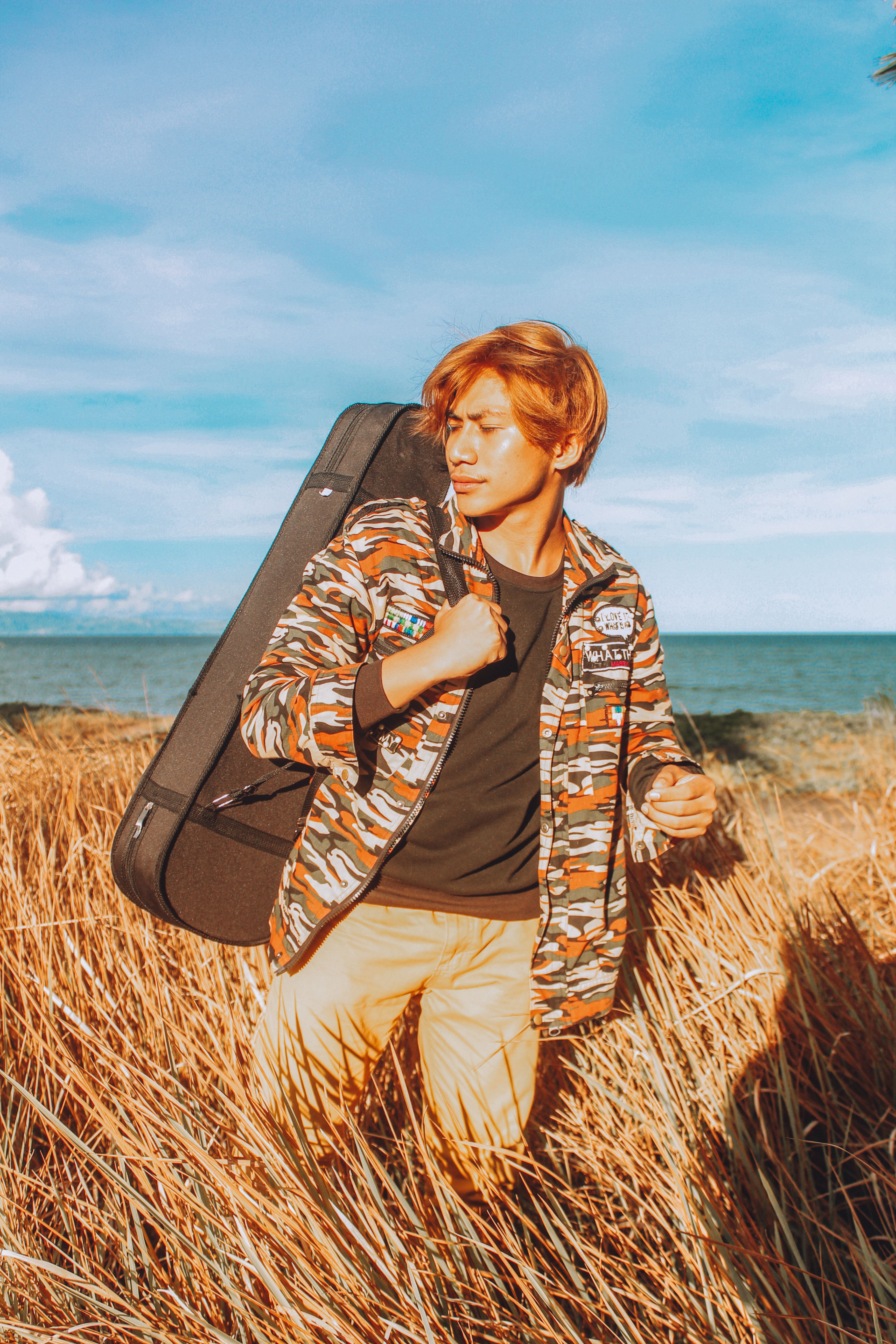 Young man in camouflage jacket carrying guitar case on sunny beach with tall grass.