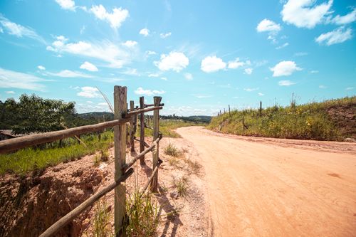 Free Scenic rural landscape featuring a dirt road and wooden fence under a bright blue sky with clouds. Stock Photo