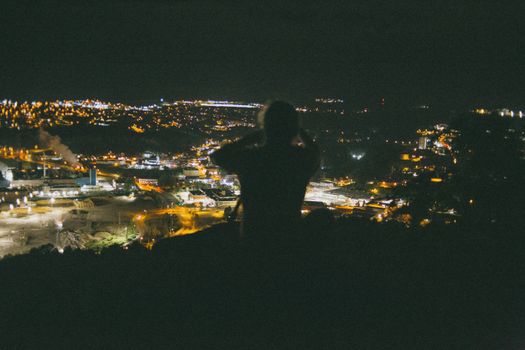 A silhouetted figure admires a bright cityscape from a dark vantage point at night.