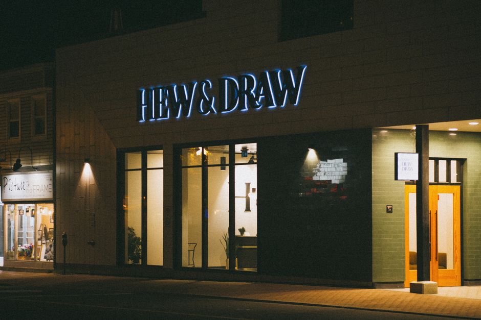 A vibrant night view of the Hew & Draw storefront with illuminated signage and modern design.
