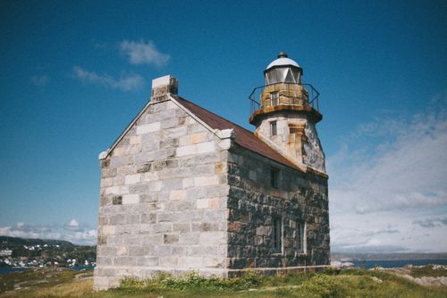 Free A vintage stone lighthouse stands against a blue sky by the calm seaside. Stock Photo