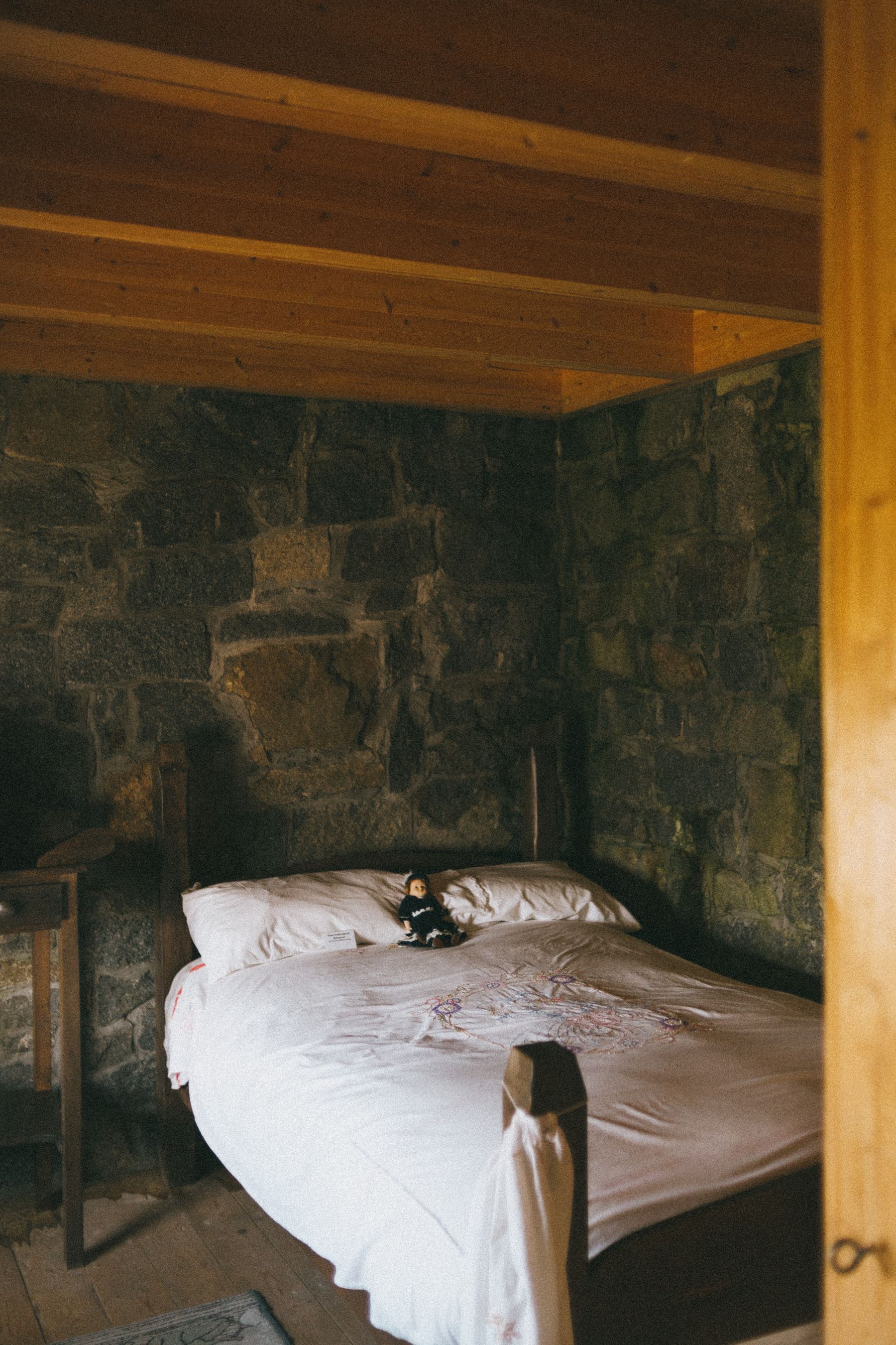 Simple bed with white linen beside a rough stone wall, timber ceiling and warm wooden floor
