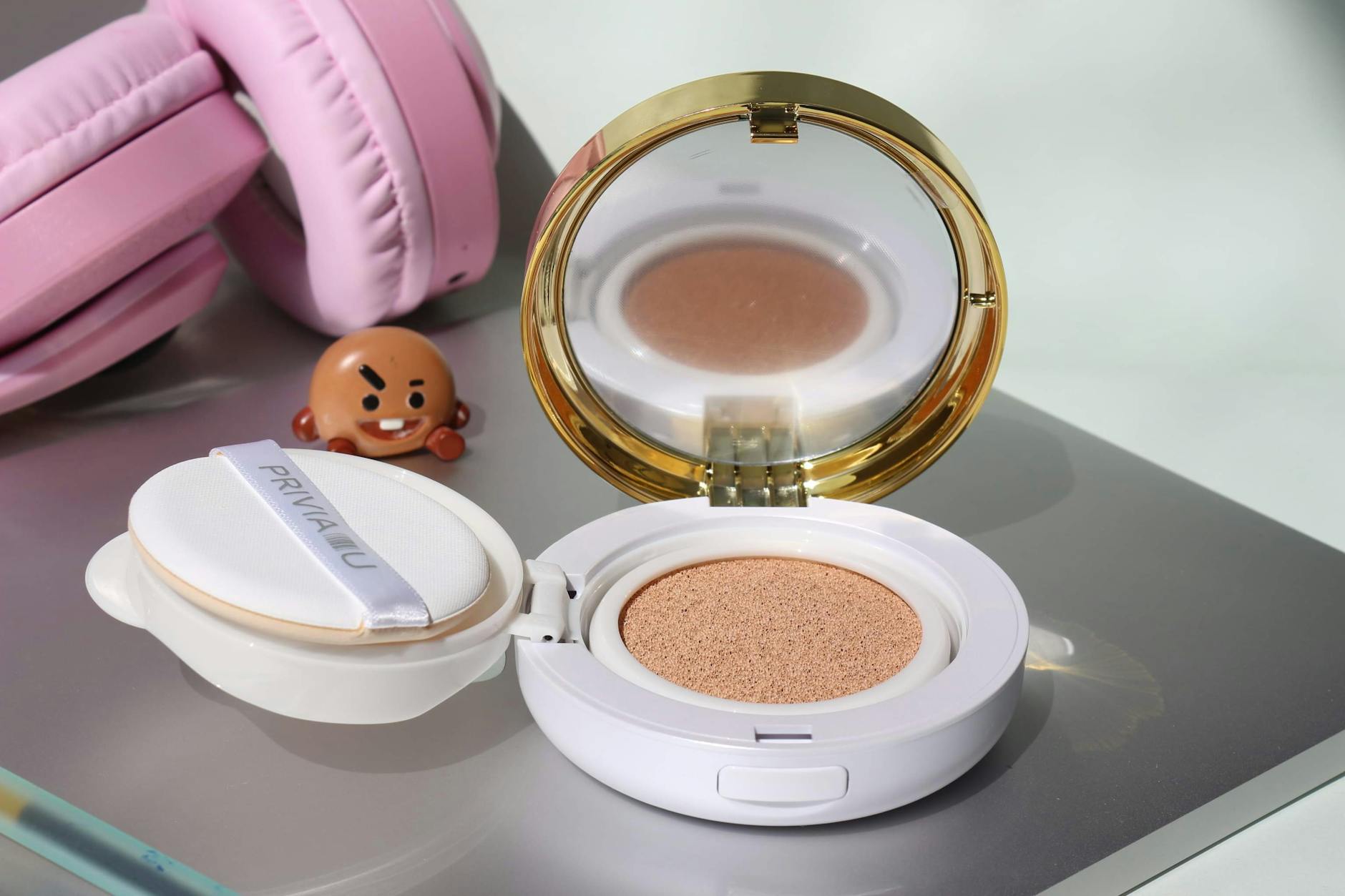Close-up of a cosmetic cushion foundation next to pink headphones and a cartoon figurine on a reflective surface.