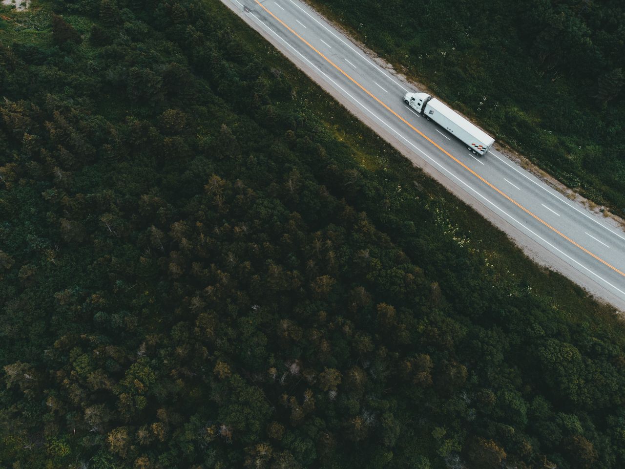 Drone shot of a container truck traveling on a forested highway during the day.