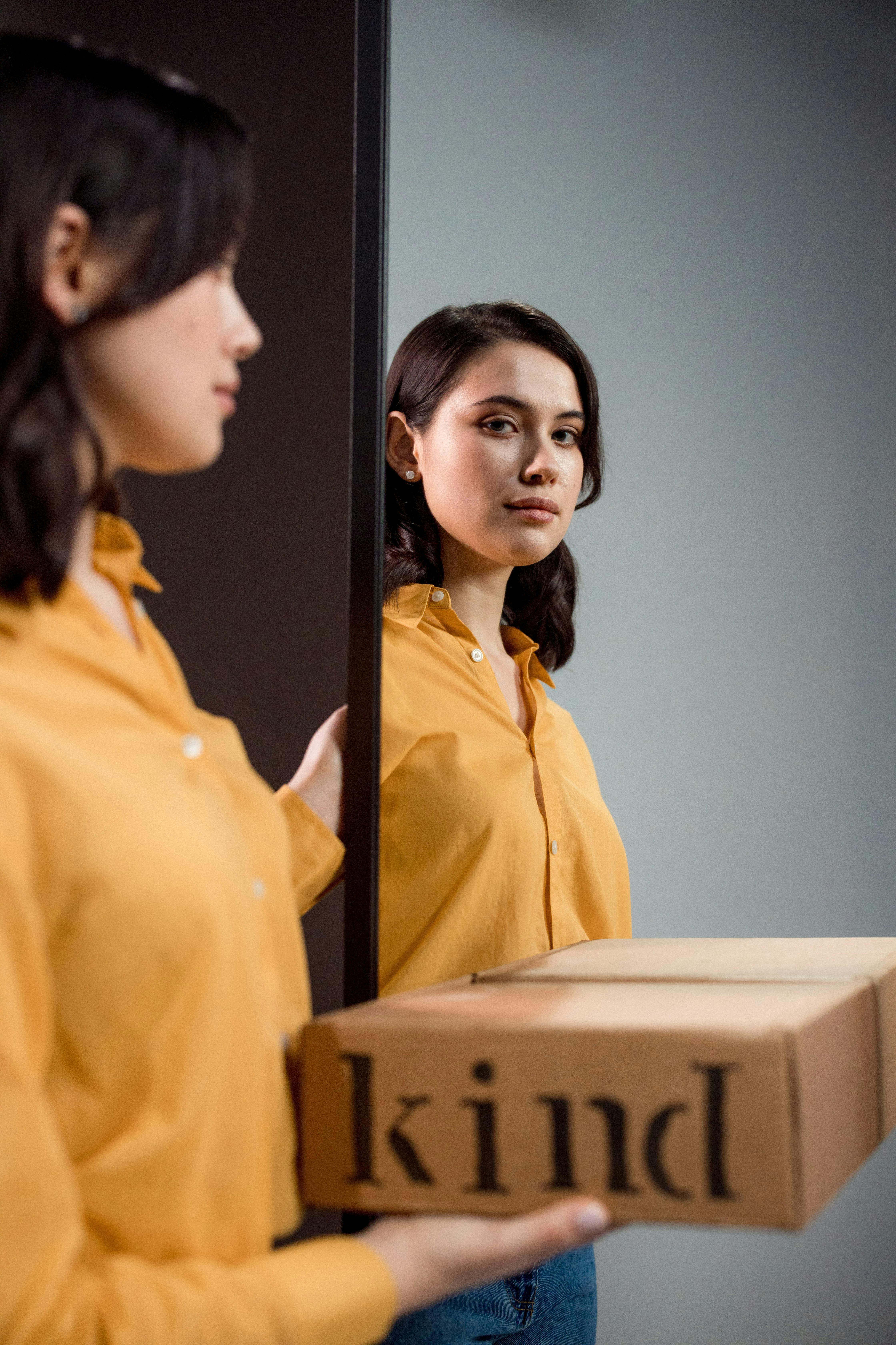 Free Woman reflects in a mirror while holding a box with the word 'kind', symbolizing inner reflection and kindness. Stock Photo